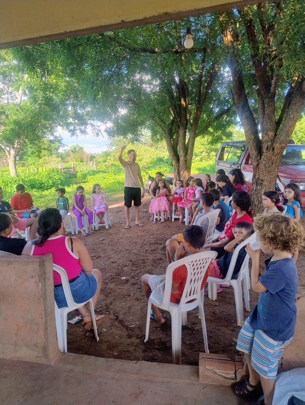 Roda de crianças e famílias em cadeiras ao ar livre, com Luan ensinando ao centro