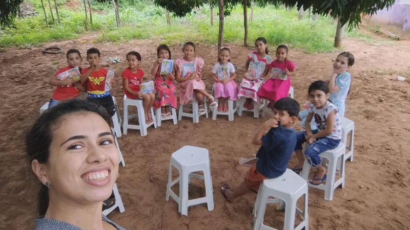 Débora sorrindo em selfie com crianças sentadas em banquinhos ao ar livre
