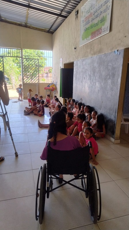 Crianças sentadas no chão ouvindo, ao lado de educadora em cadeira de rodas